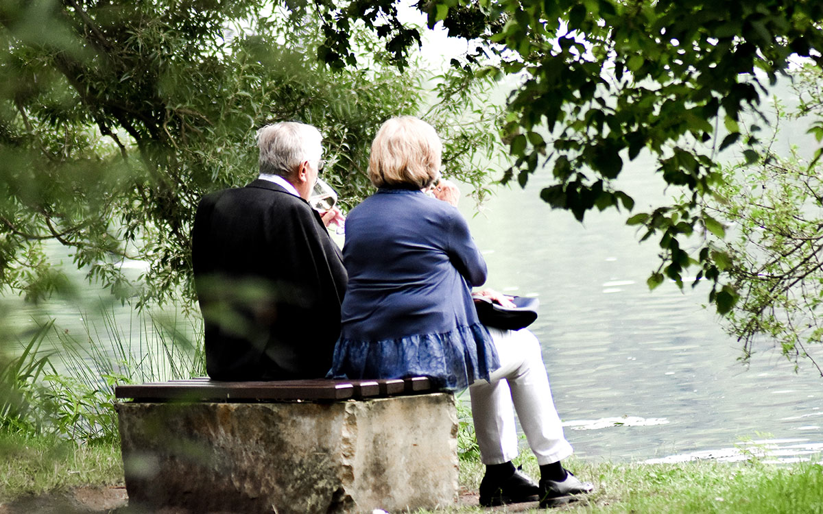 An elderly couple sits together on a bench outside.