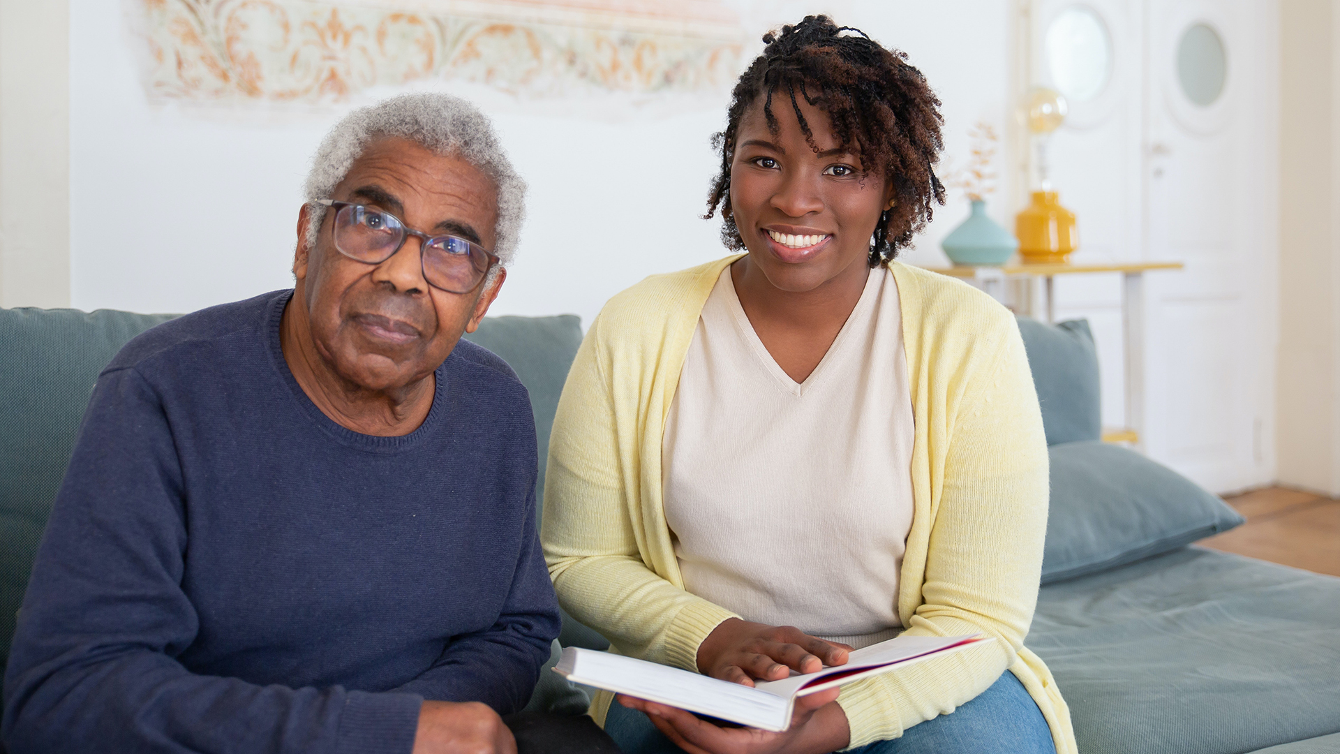 A health care aide reading to an elderly man on a couch in his home