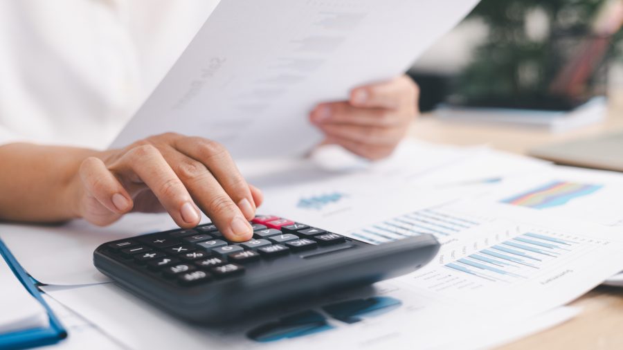 Detail of a person's hand typing on a calculator on a desk covered in papers